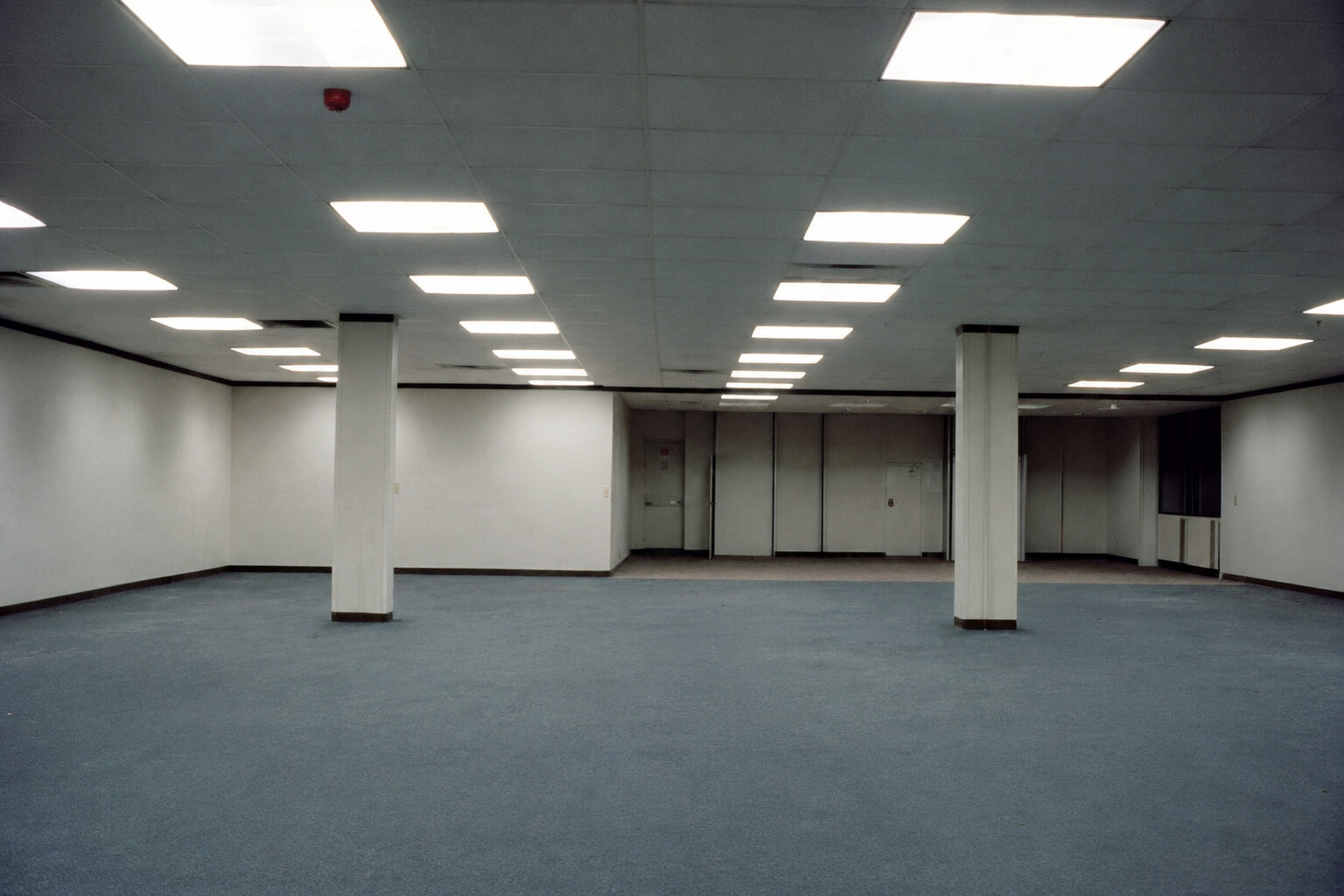 Vacant open-plan interior with fluorescent ceiling lights, blue-gray carpet, and support columns